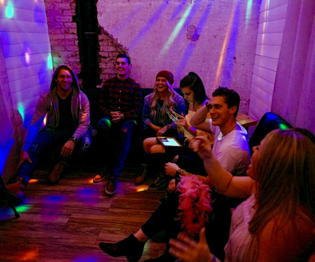 A group of young adults laughing and singing in a private karaoke room with colorful LED disco lights at Heart & Seoul Karaoke in Provo.