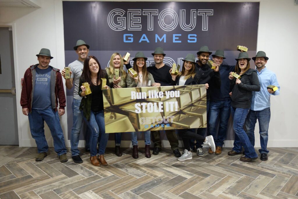 A large birthday group posing with prop gold bars and a banner that says 'Run like you stole it!' in the lobby of Getout Games escape room.
