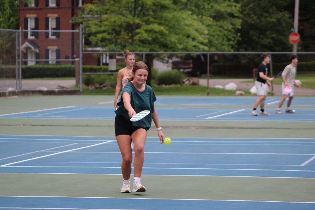 A young woman preparing to serve a yellow pickleball on a busy outdoor public court with other players visible in the background.