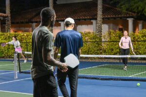 Two men holding paddles with their backs to the camera, facing a woman across the net during an evening outdoor pickleball match.