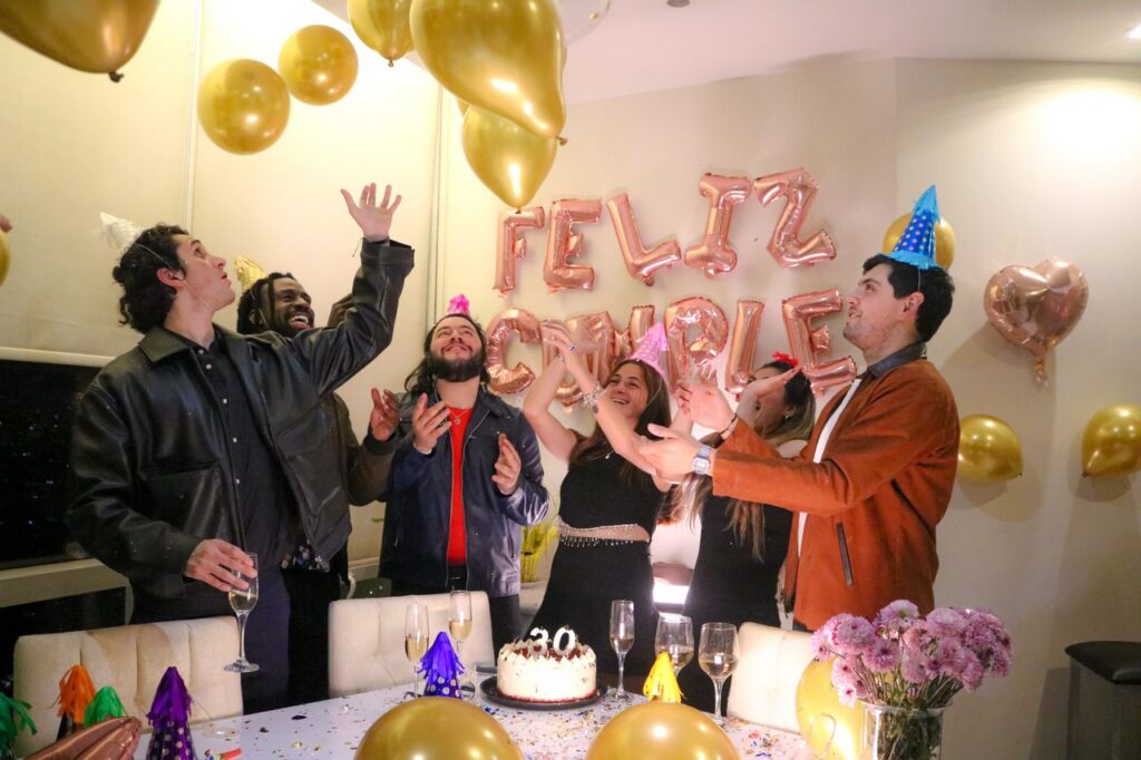 A diverse group of friends in party hats celebrating a 30th birthday with gold balloons, champagne, and a cake decorated with a 30 candle.