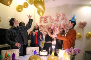 A diverse group of friends in party hats celebrating a 30th birthday with gold balloons, champagne, and a cake decorated with a 30 candle.