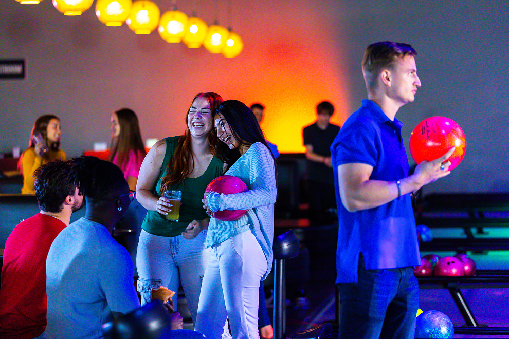 A group of young adults laughing and socializing together at a vibrant, neon-lit bowling alley during a group activity.